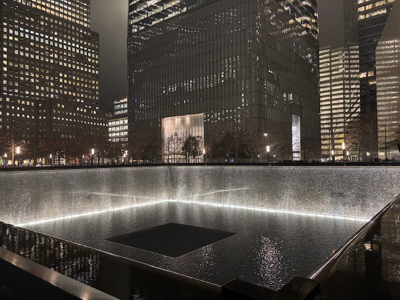 A large square hole, viewed from corner, with water falling down from the edges lit in white, with a second dark, smaller square hole in the center, surrounded by the lower floors of skyscrapers with lights in the windows, viewed at night