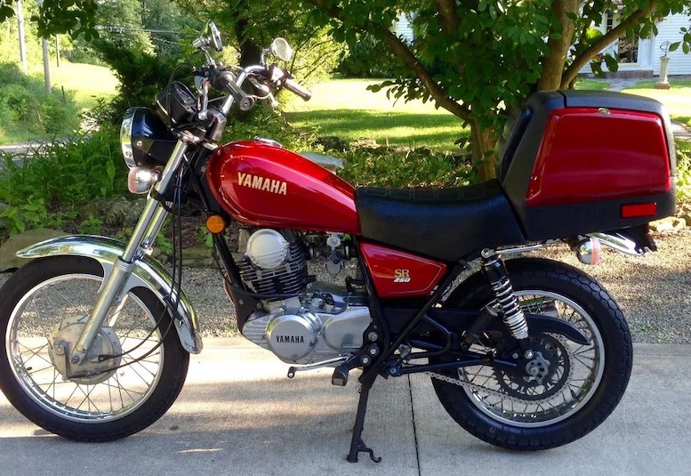 A side view from the left of a red Yamaha SR250 motorcycle with the trunk installed on the rear